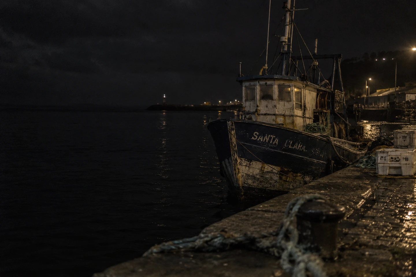 Barco de pesca atracado no cais de pedra de Porto do Funil, à noite, com luzes ao fundo.